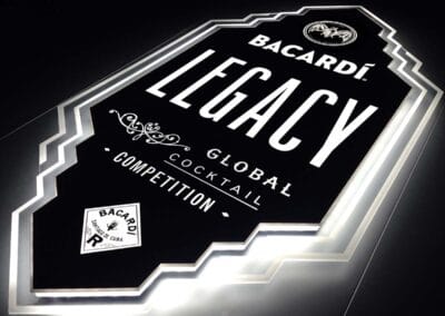 Látványos, geometrizáló, élvilágító fali display a "Bacardi LEGACY Global Cocktail Competition" felirattal és a márka logójával. Elegáns fehér fénnyel ragyog a sötét háttér előtt.