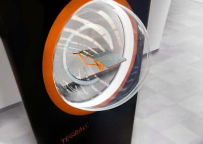 Fekete monolitszerű display, aminek a közepén narancs peremmel egy belülről kivilágított plexigömb van. A gömb közepén lebegni látszik a Teqball kisméretű modellje. Egy férfi fotózza a displayt és a modellt a háttérben.